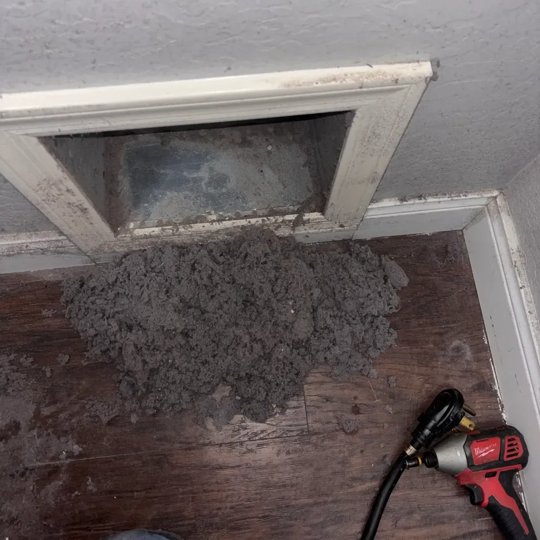Pile of dryer lint on the floor below a wall vent opening beside a drill