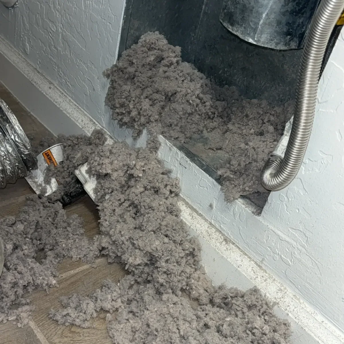 Enormous pile of gray dryer lint spilling out of a wall vent opening onto a laundry room floor